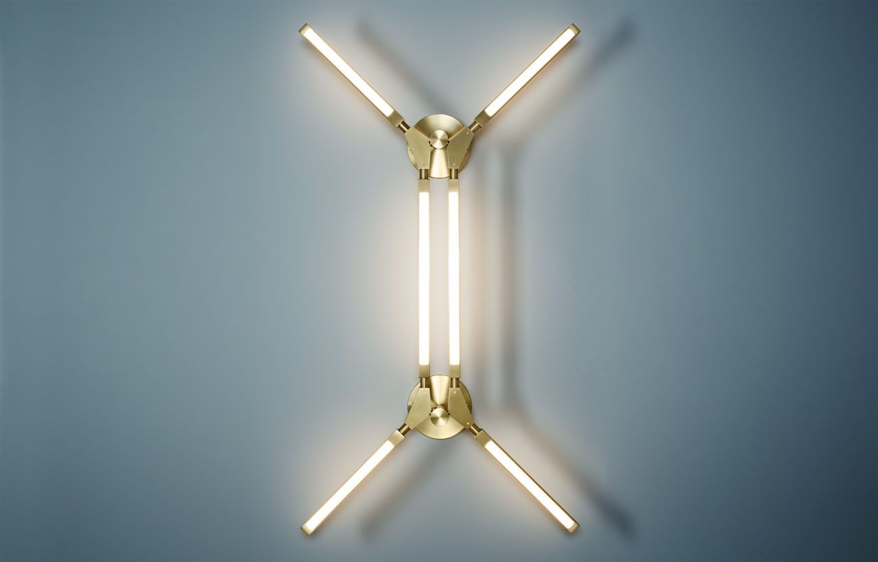 Front view of an illuminated Minor sconce with satin brass metal components on a soft blue wall.