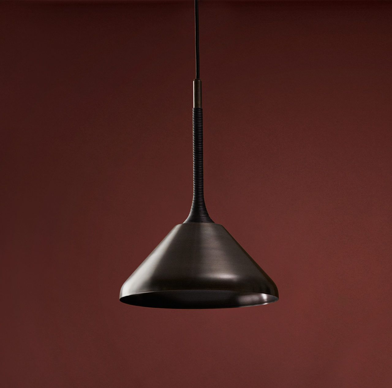 Pelle’s Stem Shade 12 in black brass with black leather coiling hangs against a deep red background.