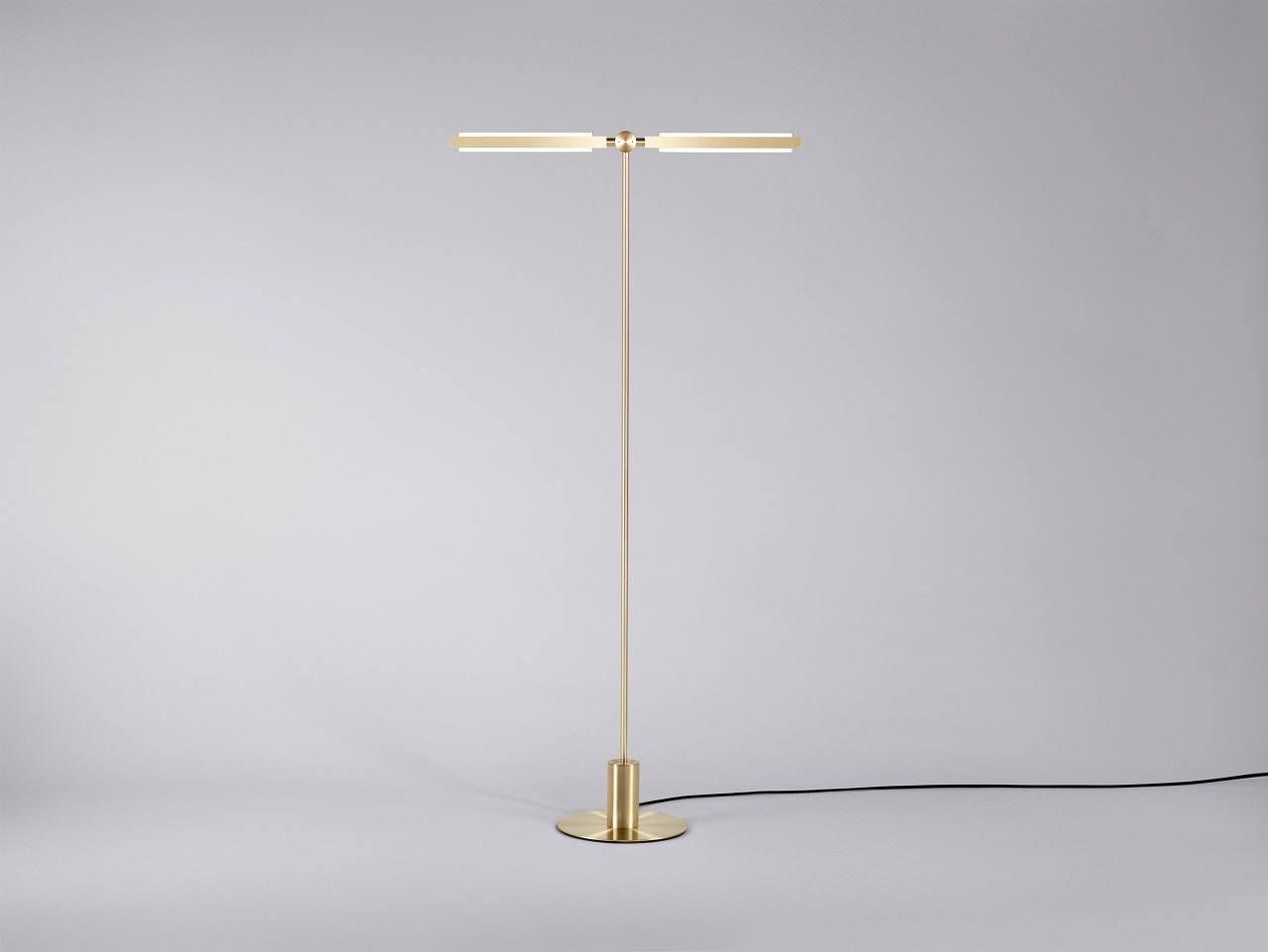 Front view of an illuminated Pris T floor lamp with satin brass metal components.