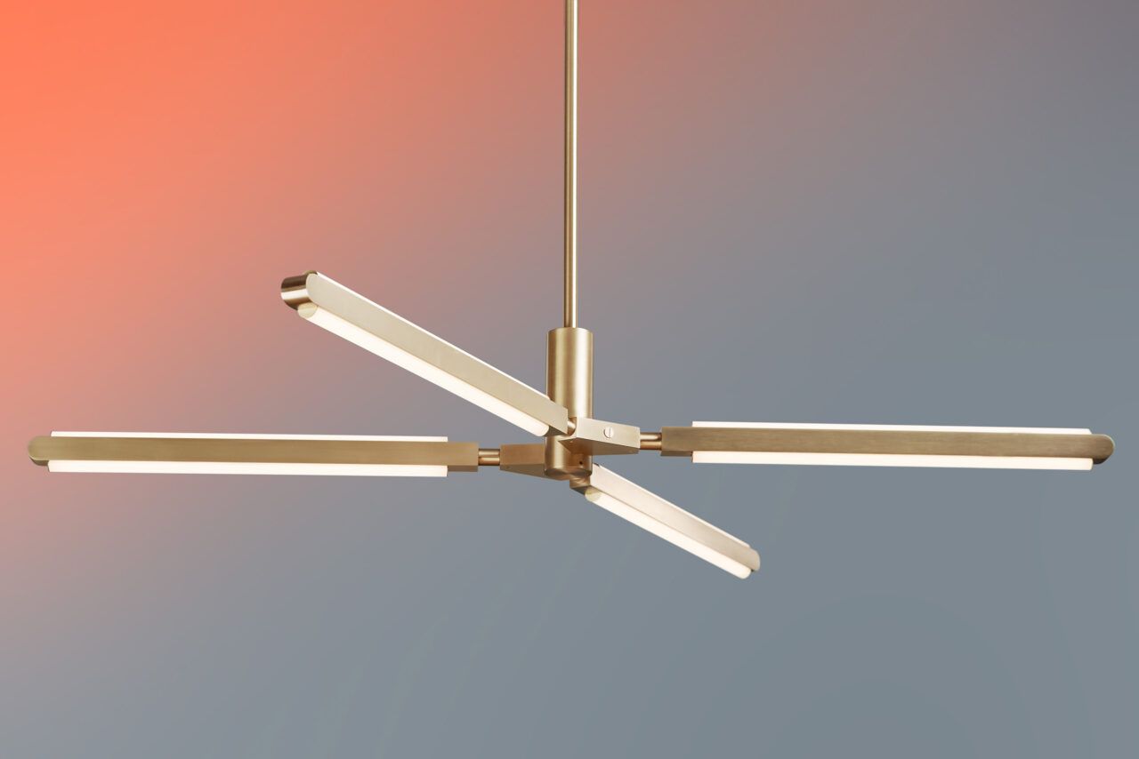 Three quarter view of an illuminated Pris X chandelier with satin brass metal components in front of a orange red to blue gray gradient.