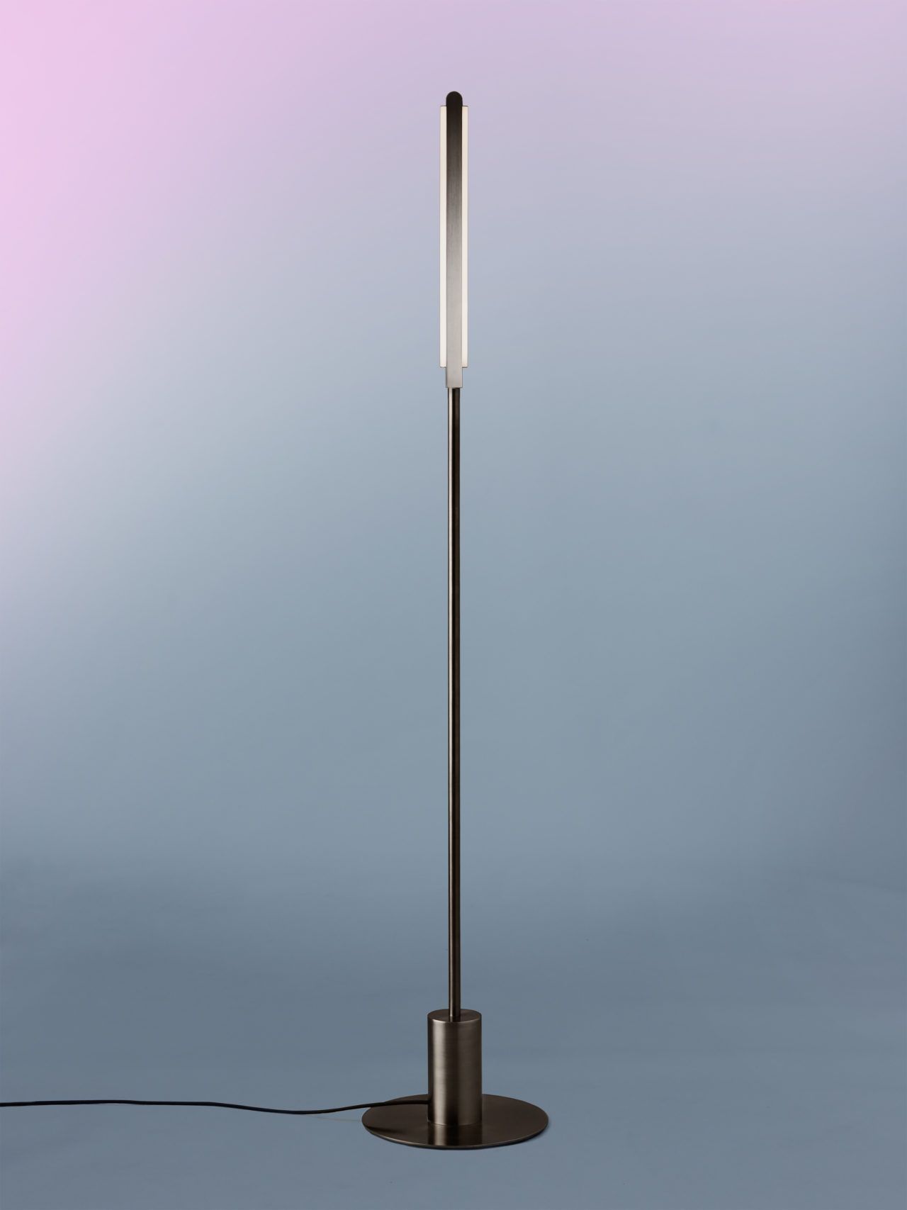 Side view of an illuminated Pris I floor lamp with black brass metal components in front of a lavender to blue gray gradient.